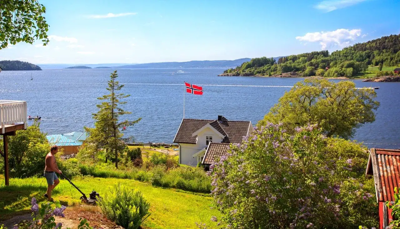 Man klipper gräset vid ett hus med norsk flagga vid en vacker fjord. En idyllisk sommardag i son Norge.