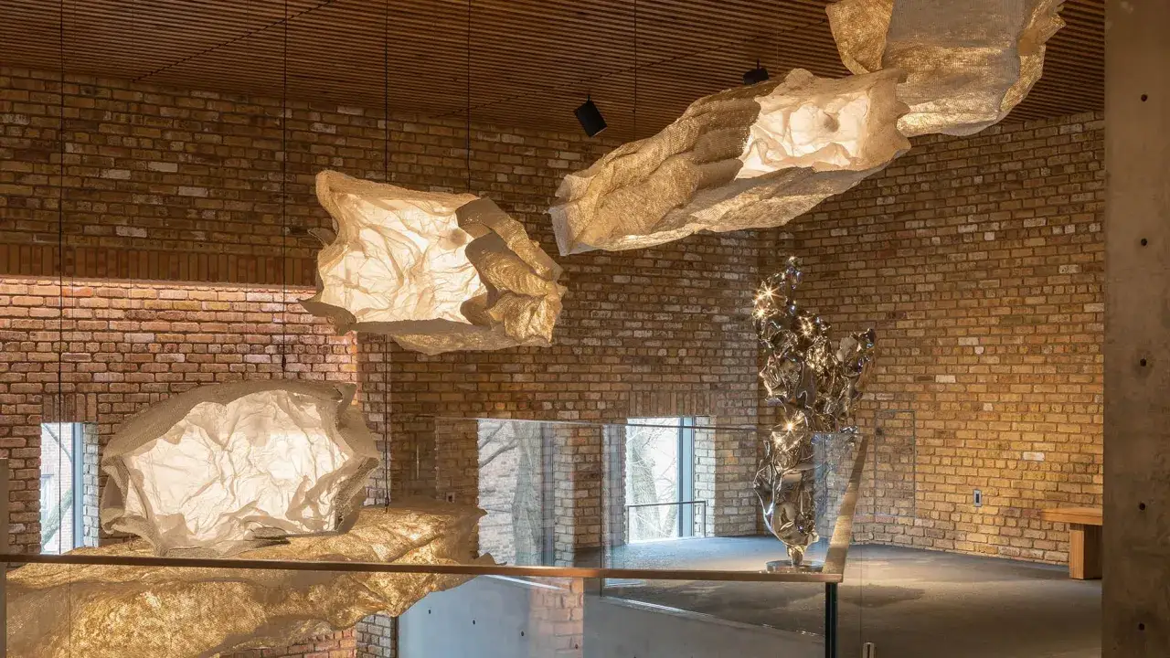 Illuminated, cloud-like sculptures hang in a space with brick walls, reminiscent of the innovative spirit of chinese contemporary artists.
