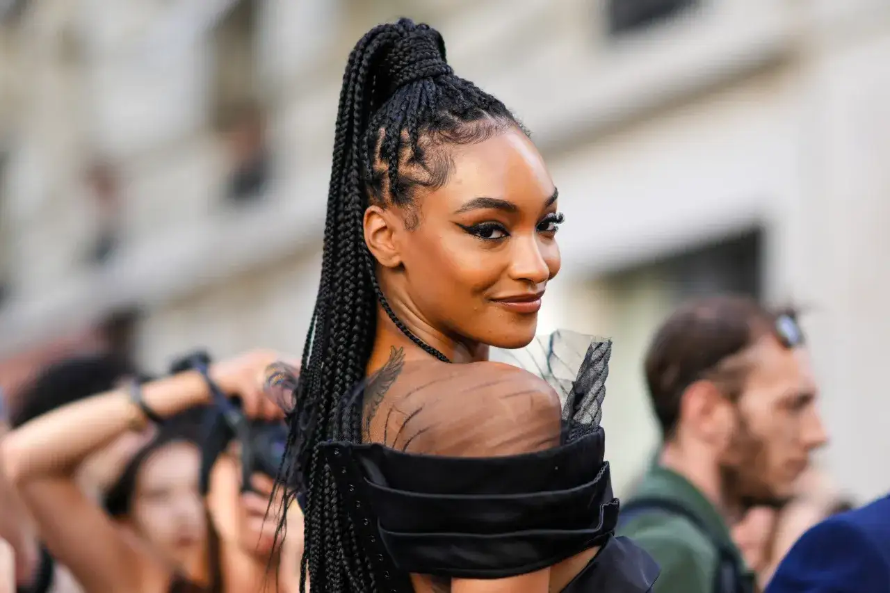 Mujer con trenzas de moda recogidas en una coleta alta, luciendo un vestido negro con detalles de tul y un tatuaje en el hombro.