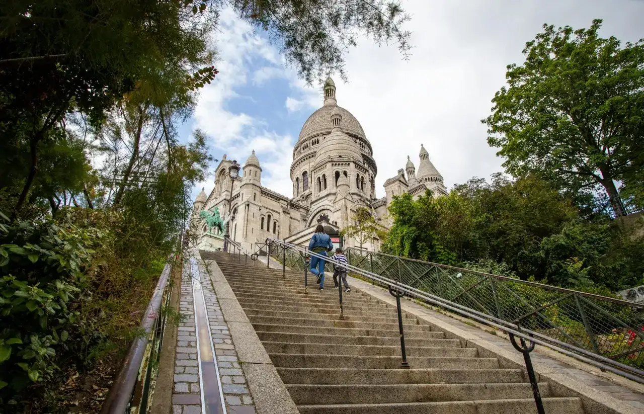 Dwie osoby wspinają się po schodach na wzgórze Montmartre, w tle Bazylika Sacré-Cœur.