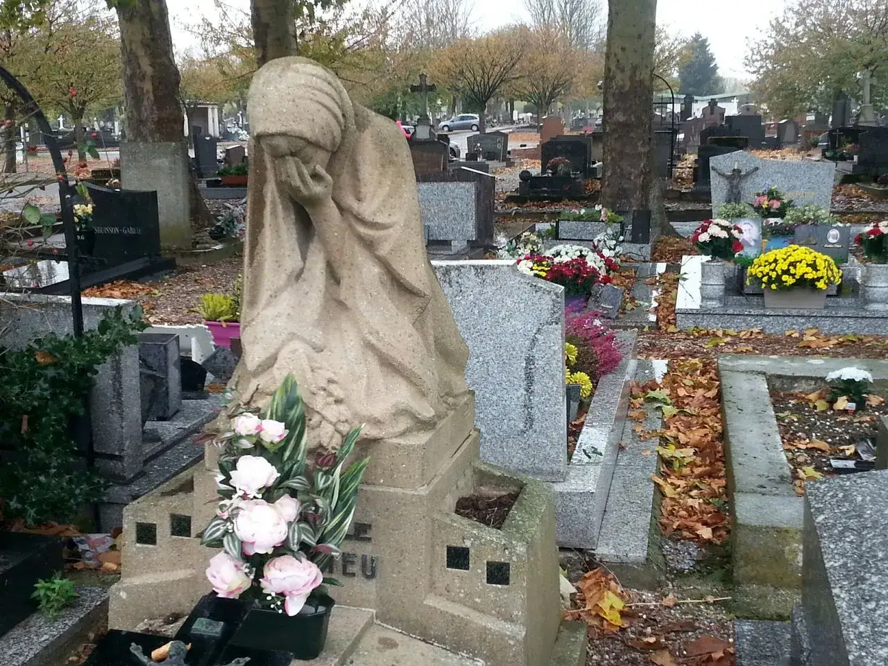 Sculpture d'une femme en pleurs au cimeti&egrave;re du sud Nancy, entour&eacute;e de fleurs et de feuilles d'automne.