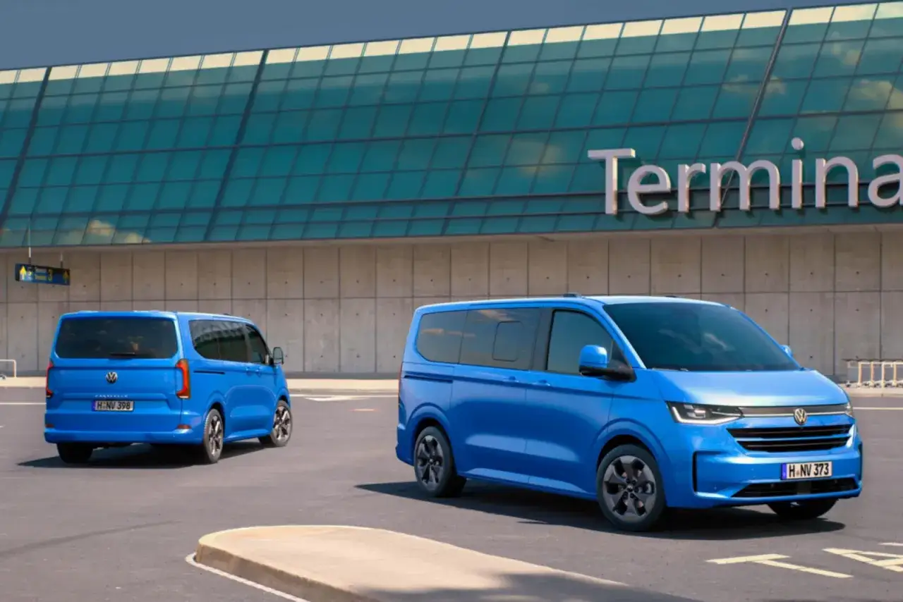 Dwa niebieskie VW Transporter T7 zaparkowane przed budynkiem terminala lotniska.