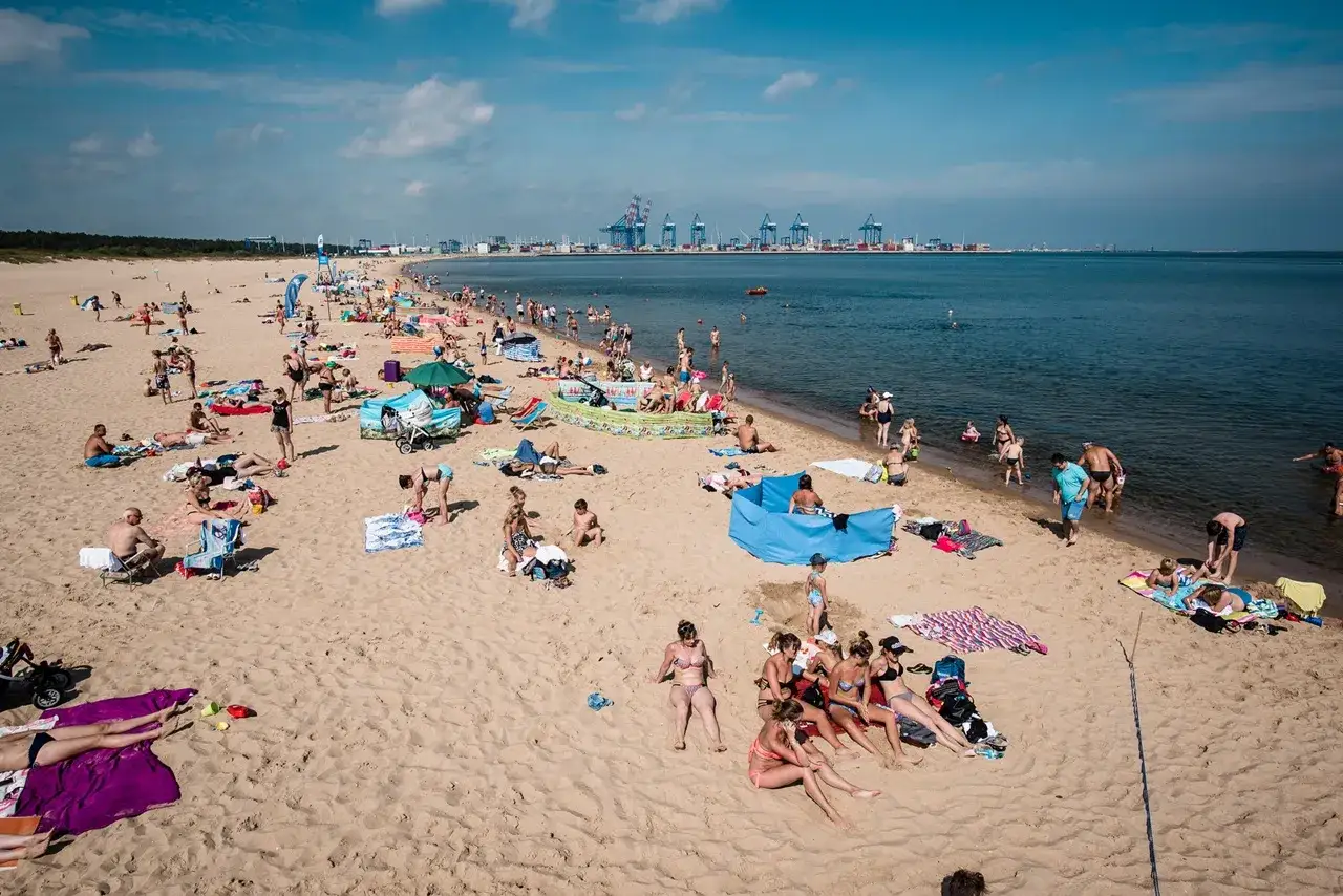 Tłum ludzi wypoczywa na słonecznej plaży Stogi. W tle widać portowe dźwigi i morze.