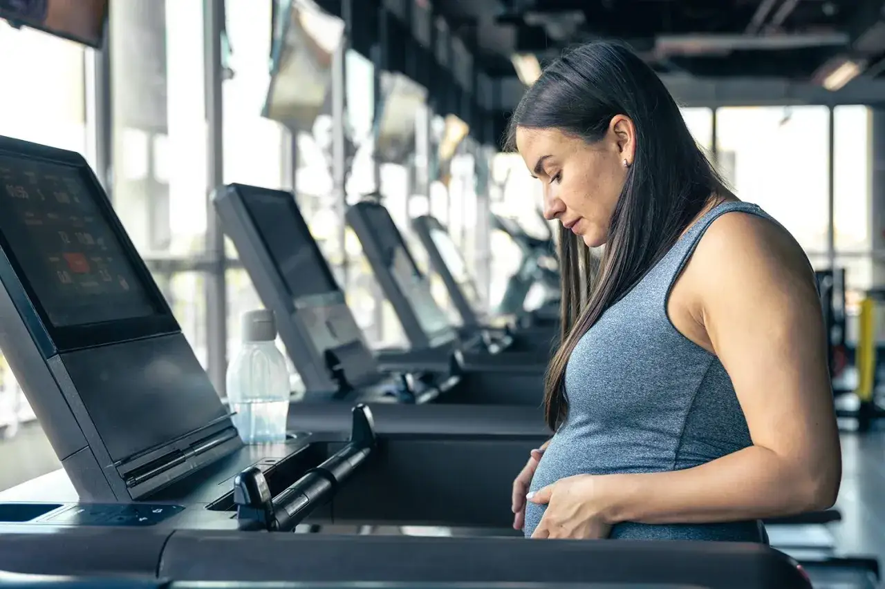 Donna incinta accarezza il pancione mentre si allena su un tapis roulant in palestra.