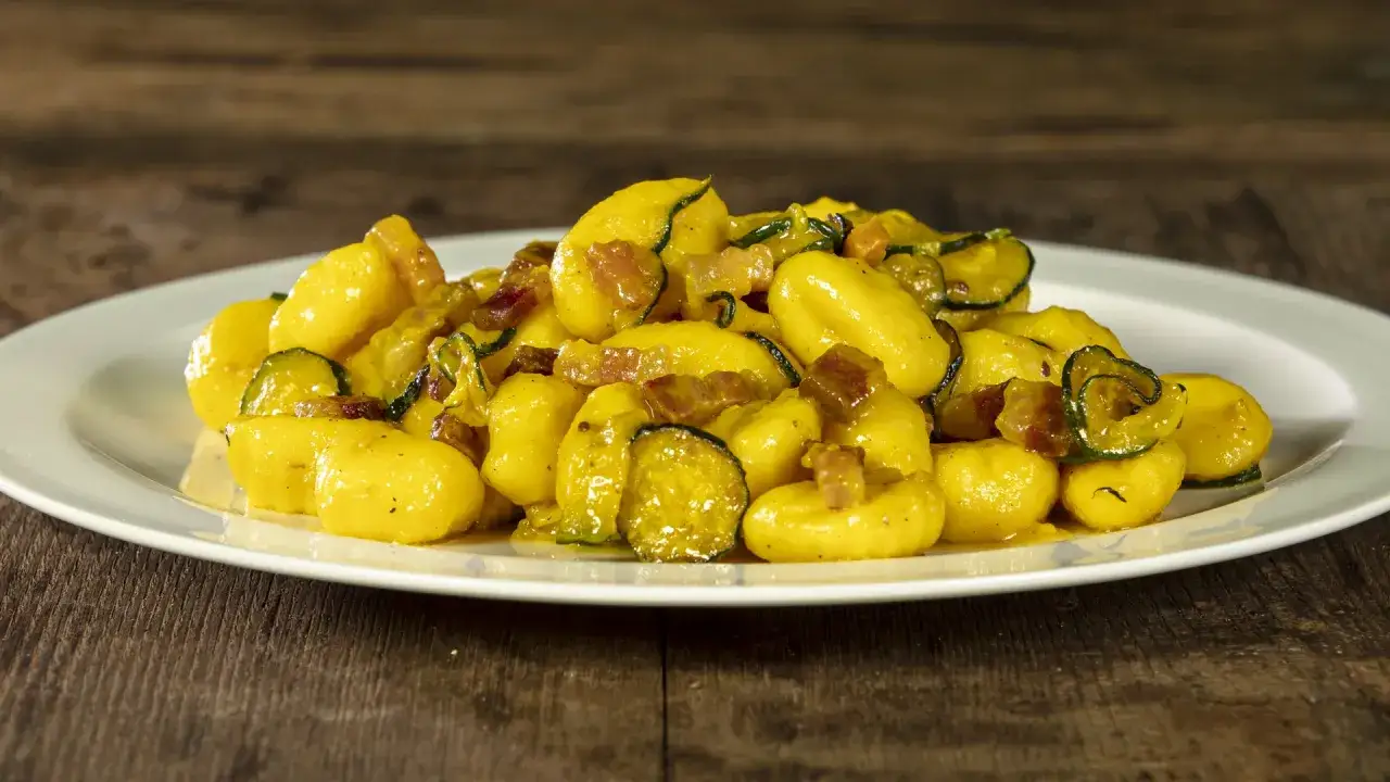 Gnocchi di zucchine dorati e saporiti, conditi con zucchine a rondelle e cubetti di pancetta croccante, serviti su un piatto bianco.