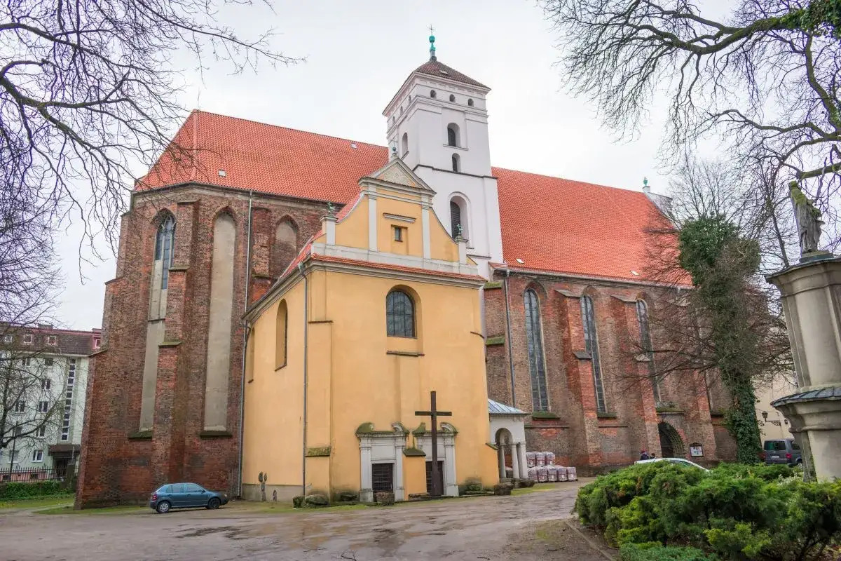 Kościół Bożego Ciała z cegły i żółtymi elementami, z wieżą i czerwoną dachem, otoczony drzewami.