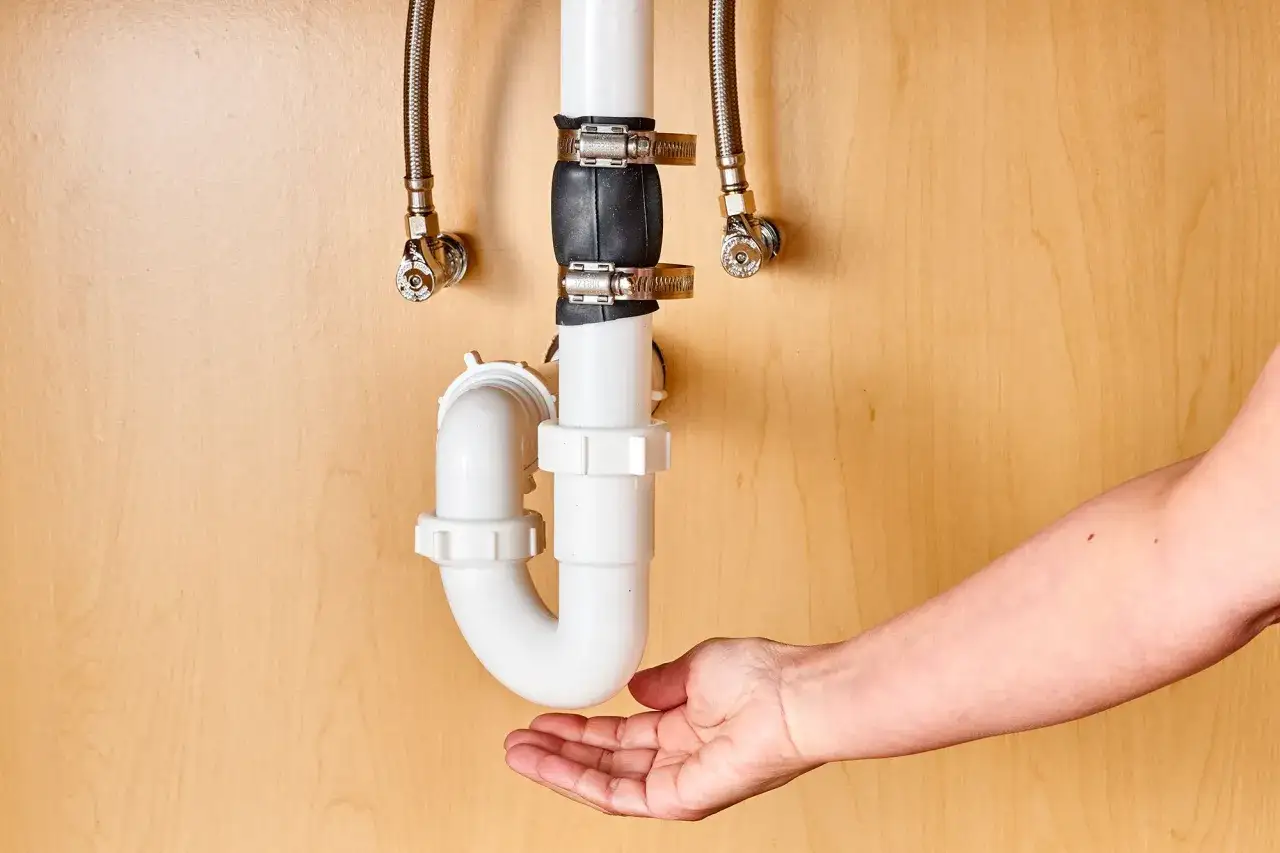 A hand is shown beneath a P-trap, demonstrating how to cut PVC pipe for plumbing repairs.