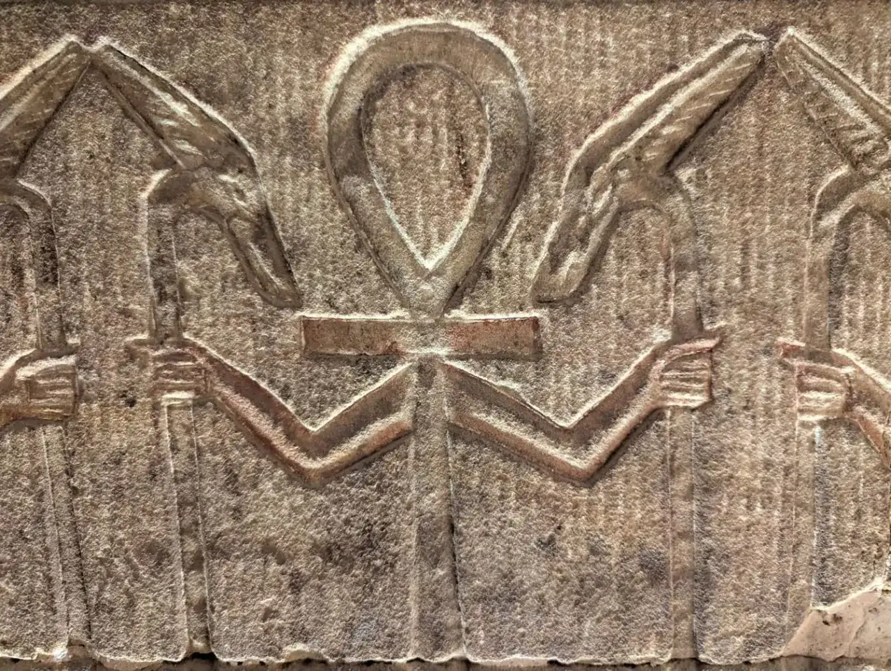 Starożytny relief z symbolem ankh, trzymanym przez postacie z głowami zwierząt.