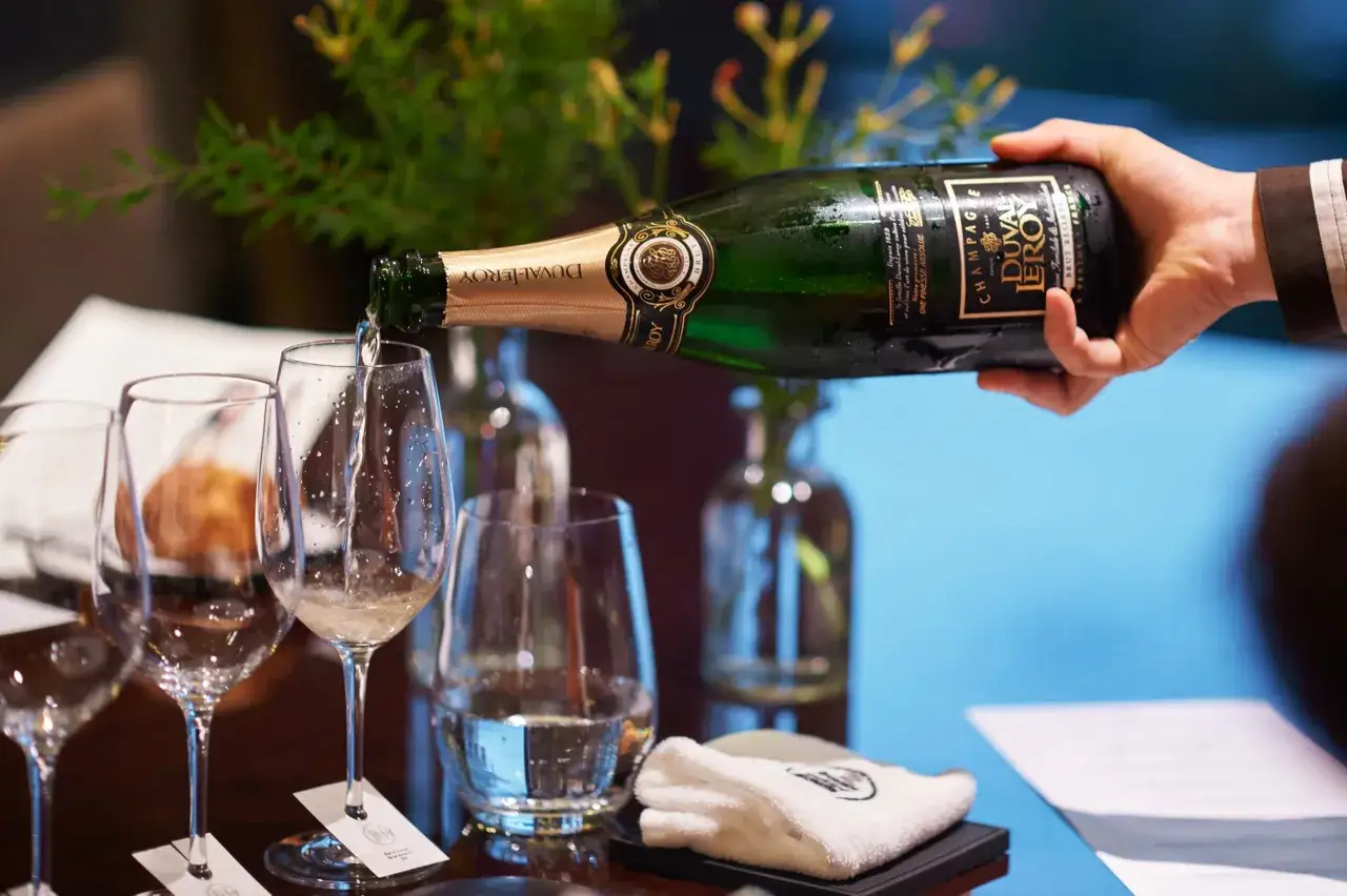 Une main verse du champagne d'une bouteille Duval Leroy dans des verres. Une bouteille de 9 litres est tenue avec soin.