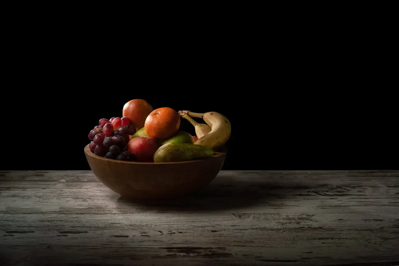Un cesto di frutta in un shooting still life: uva, arancia, mele, pere e banane su un tavolo di legno scuro.
