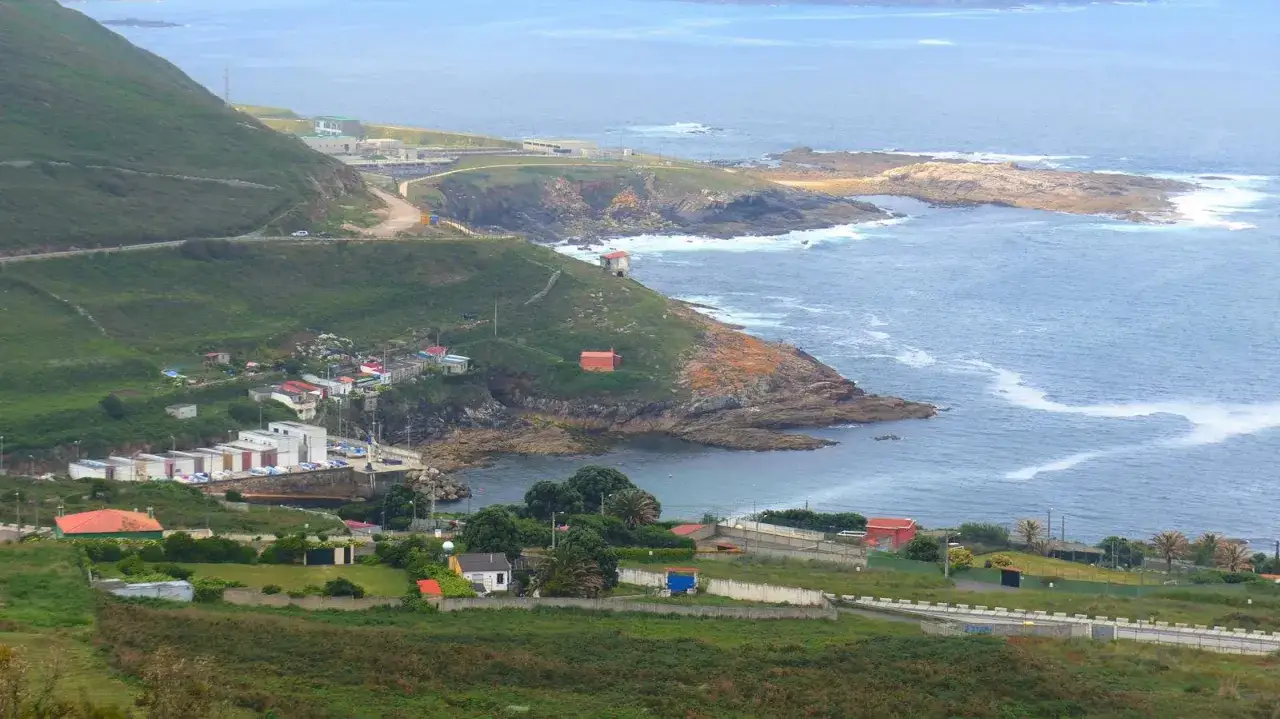 Vista de un peque&ntilde;o puerto pesquero, o porti&ntilde;o, con casas y edificios industriales en una ladera verde junto al mar.
