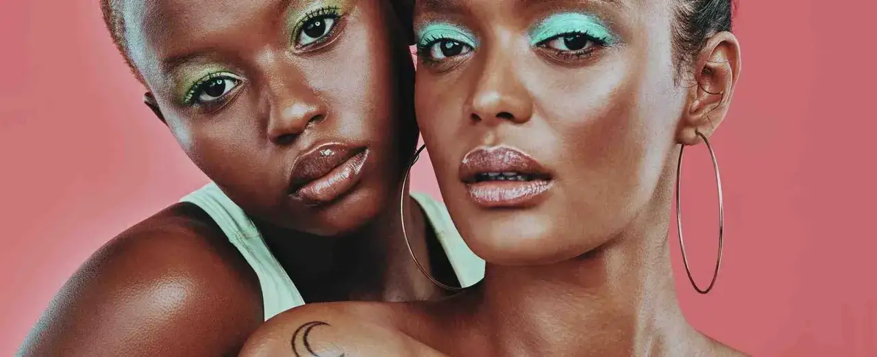 Dos mujeres con maquillaje vibrante. Una tiene sombra de ojos verde menta y la otra azul turquesa, como una **línea de agua** que resalta su mirada.