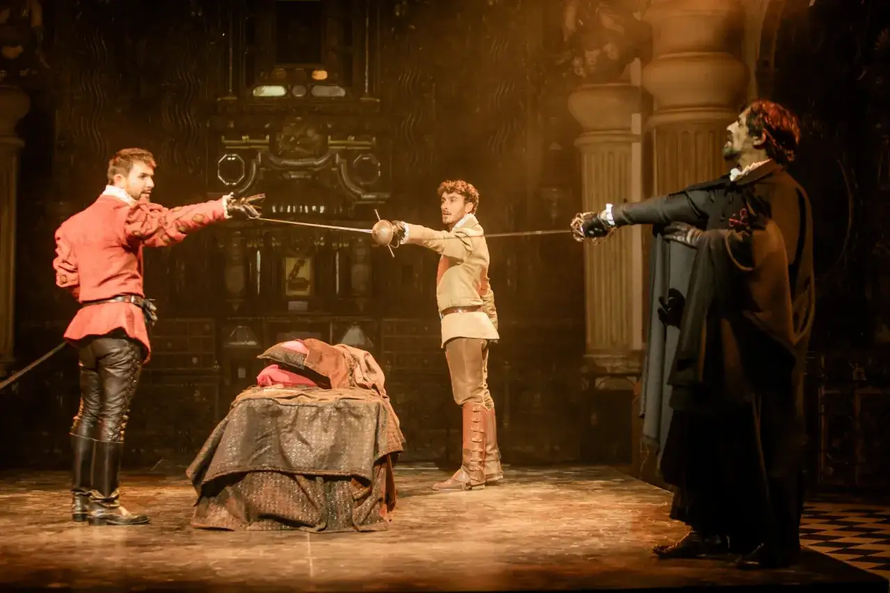 Escena de duelo de espadas, un resumen de Don Juan Tenorio. Tres hombres con trajes de época se enfrentan en un escenario teatral.