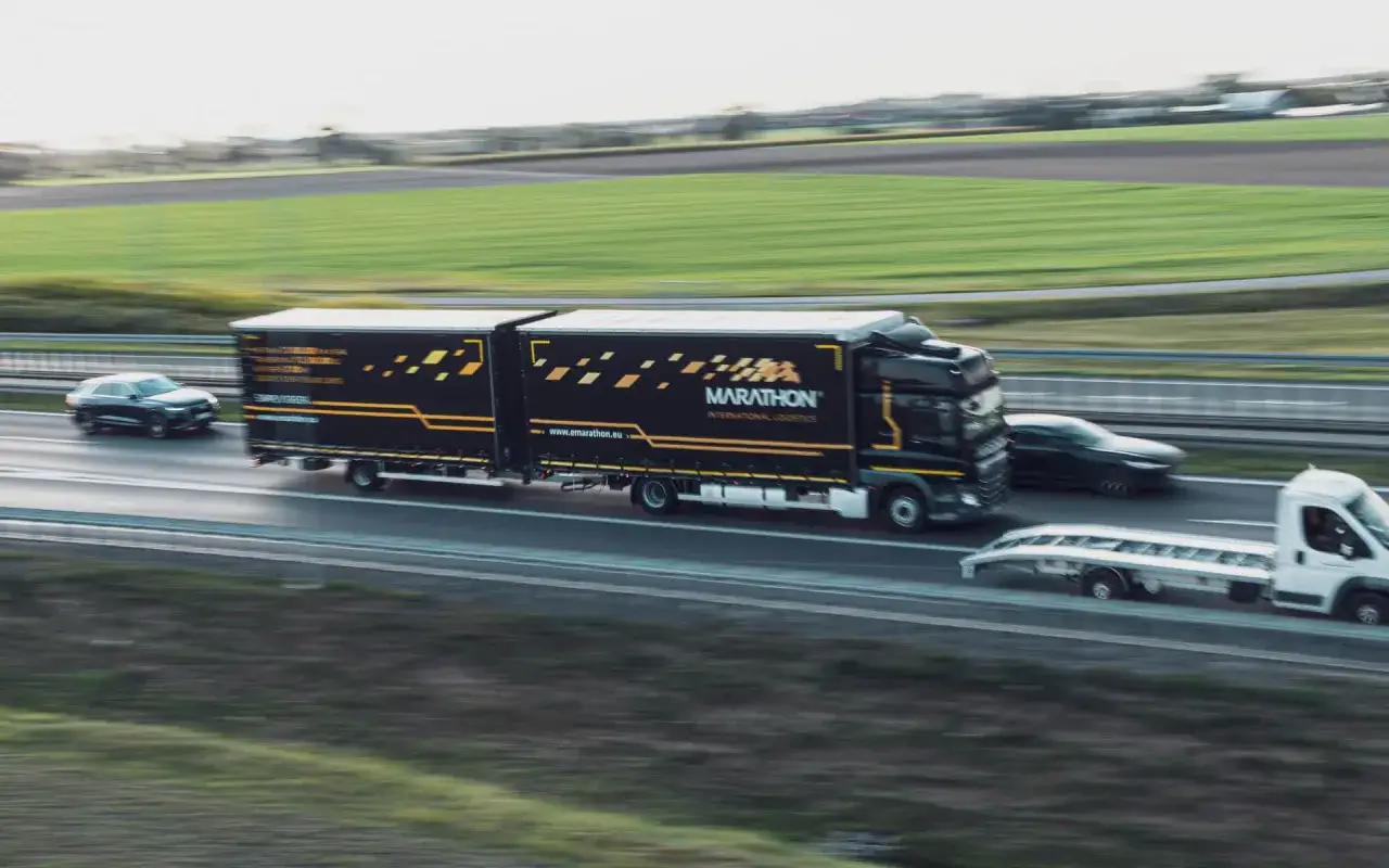 Ciężarówka Marathon International Logistics na autostradzie, obok samochód osobowy. Efektywne delegowanie kierowców to klucz do sprawnego transportu.