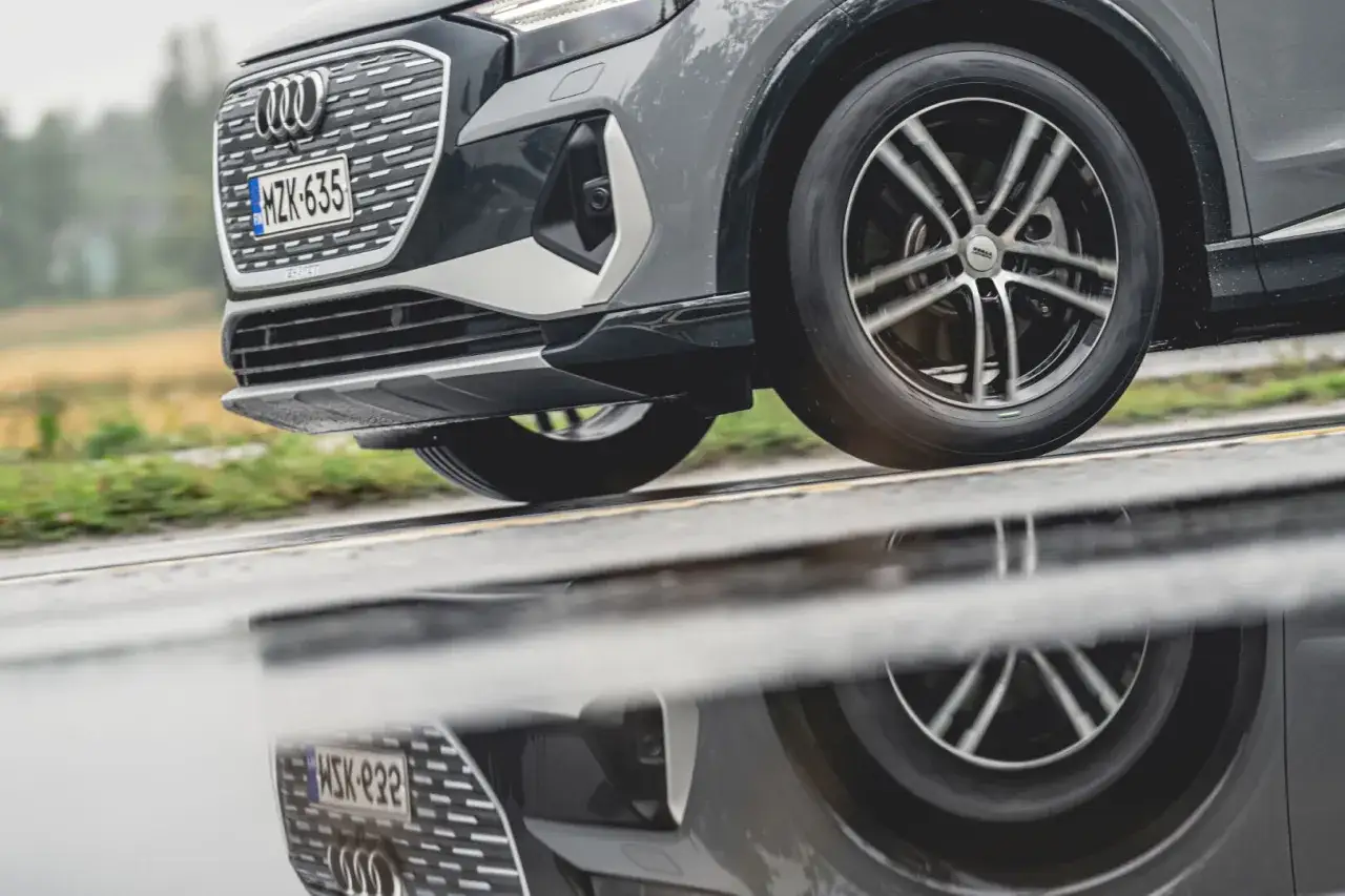 En Audi kör på en regnig väg, dess hjulbultar syns tydligt i den blöta asfalten som reflekterar bilen.