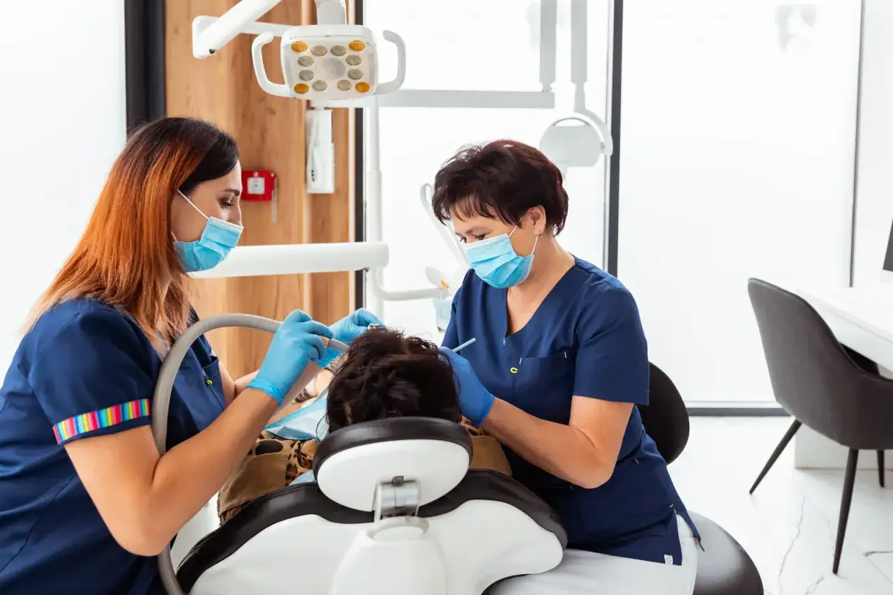 Dwie dentystki w maseczkach i rękawiczkach wykonują zabieg u pacjenta siedzącego na fotelu dentystycznym.
