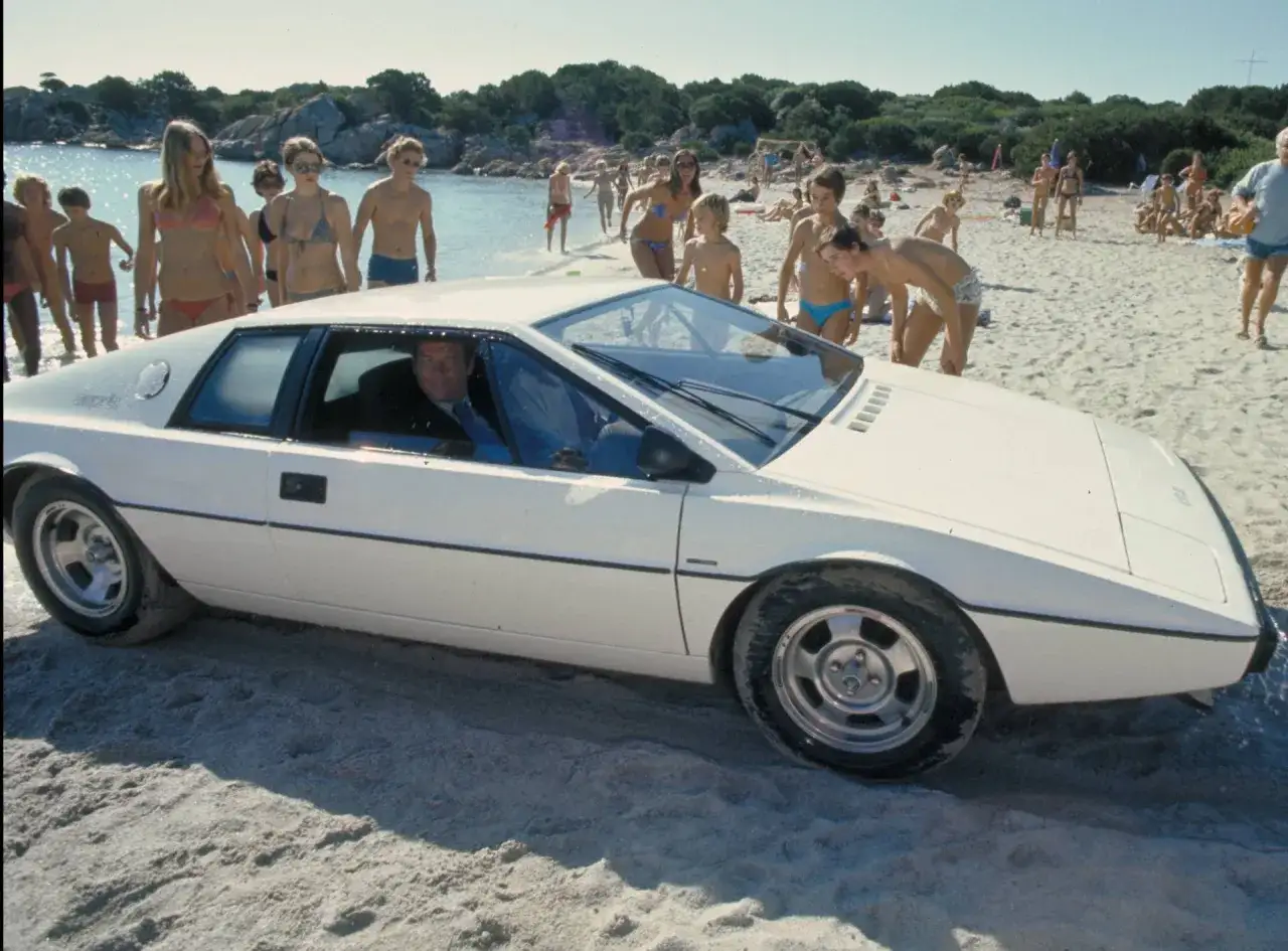 James Bond k&ouml;r sin ikoniska vita Lotus Esprit p&aring; en solig strand, omgiven av badande m&auml;nniskor.