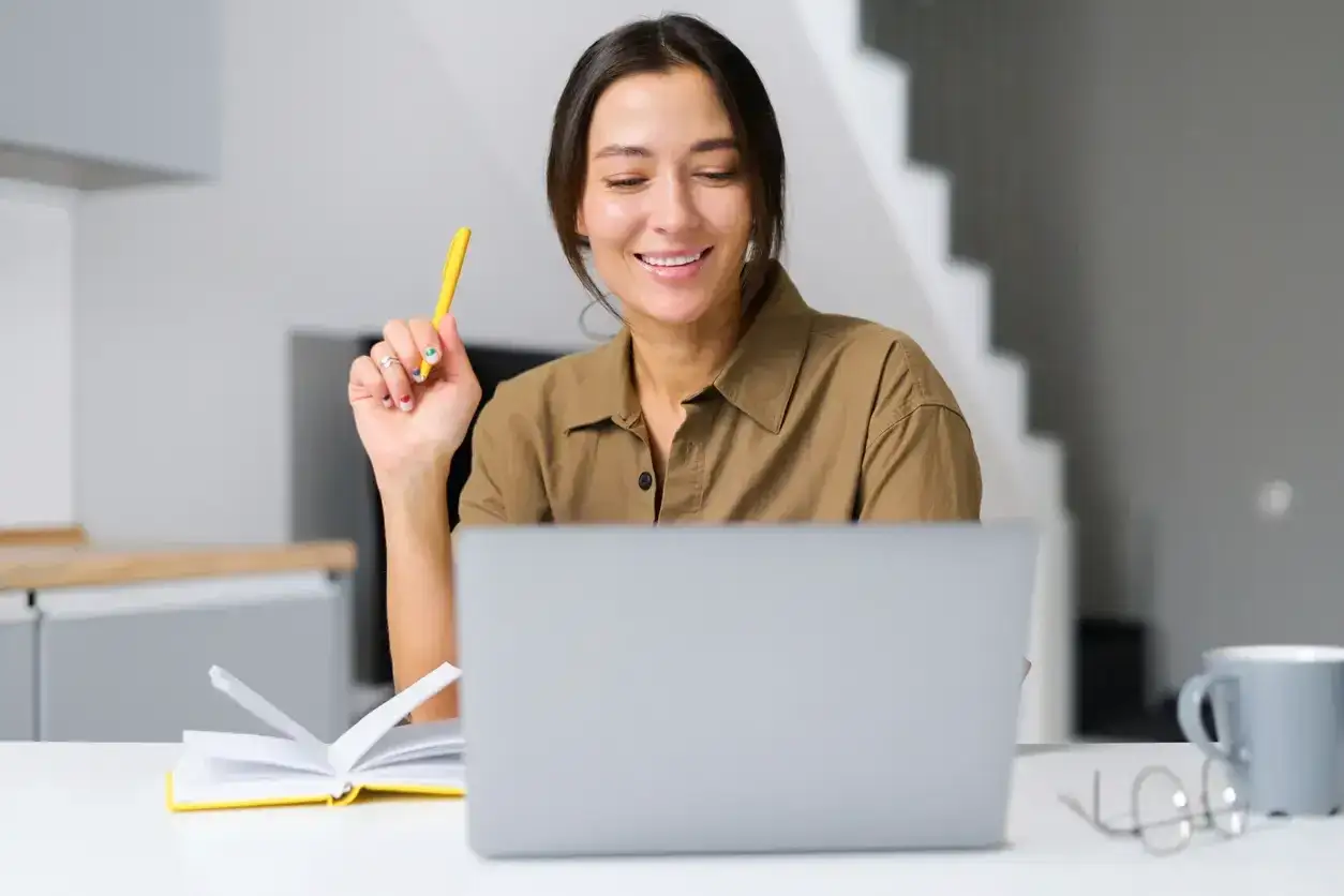 Lächelnde Frau mit Stift am Laptop, fragt sich, ob sich ein Fernstudium lohnt. Notizbuch und Brille liegen bereit.