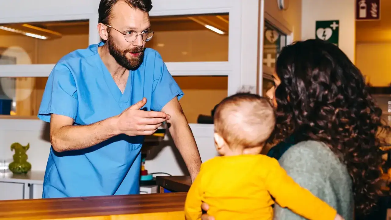 Läkare i blå scrubs pratar med en mamma som håller sitt barn. Kanske måste han avbryta semester för att hjälpa dem.