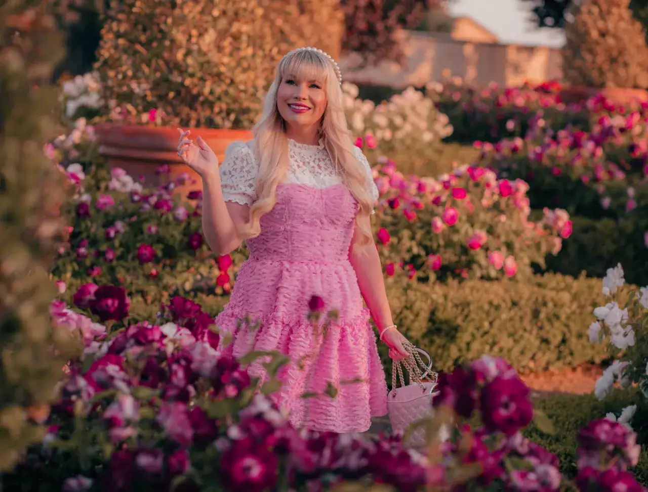 Barbie w świecie mody, w r&oacute;żowej sukience i z torebką, otoczona kwitnącymi r&oacute;żami.