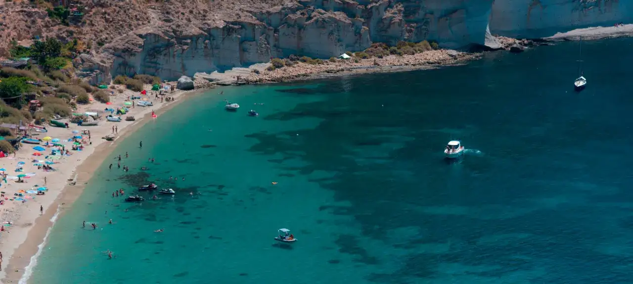 Gente disfruta del sol y el mar en la playa de San Pedro, con barcos y motos acuáticas en las aguas cristalinas.