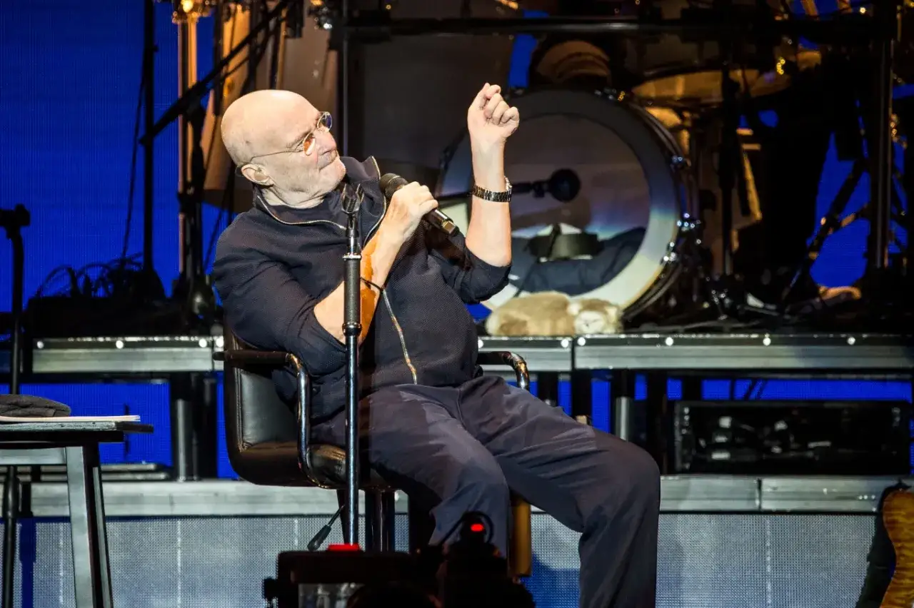 Phil Collins, mimo choroby, śpiewa na koncercie, siedząc na krześle.