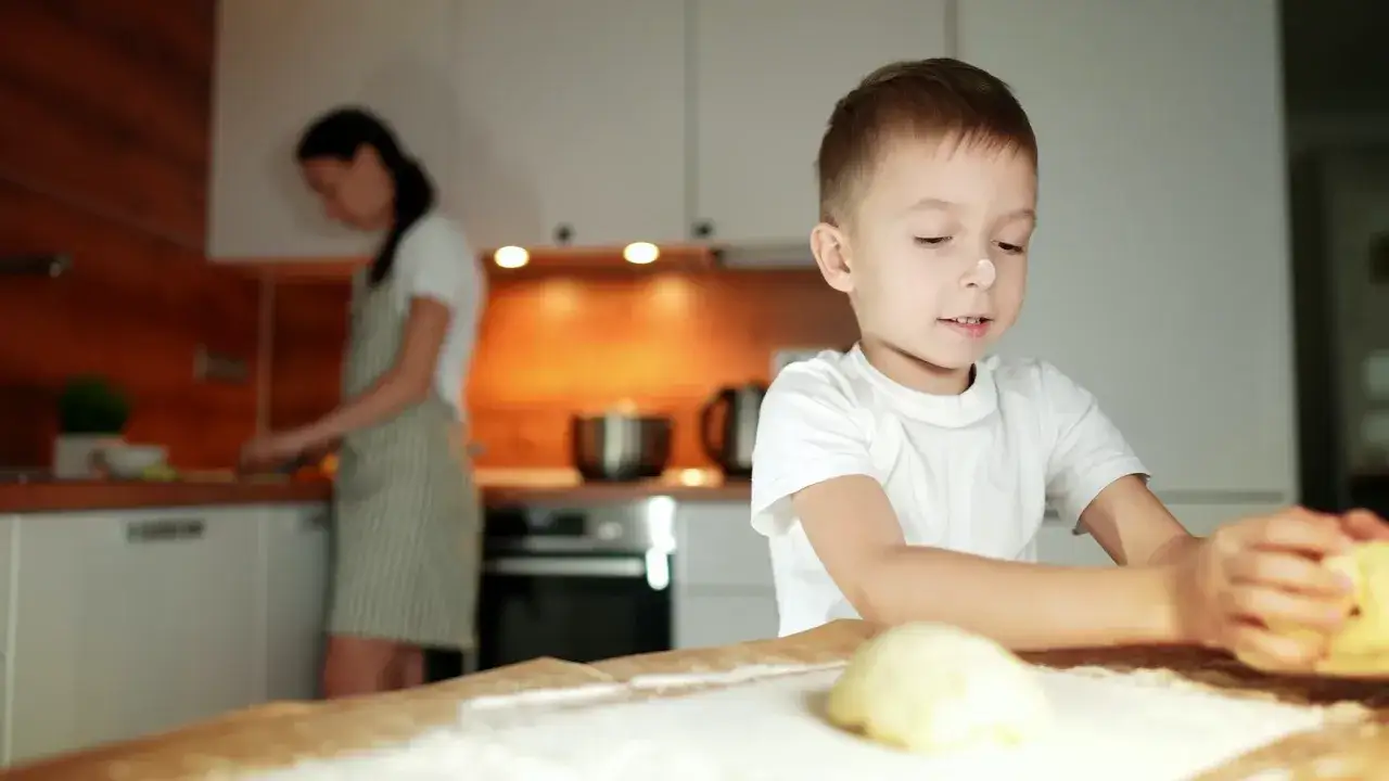 Chłopiec z mąką na nosie lepi ciasto. Mama w tle gotuje. To świetny sposób, jak motywować dziecko do nauki przez zabawę.