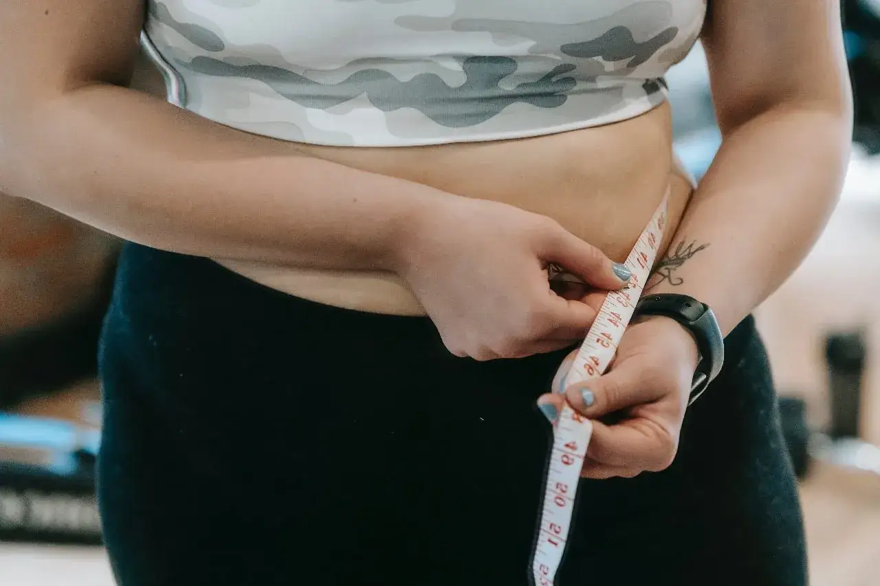 Kobieta mierzy obwód talii, kontrolując **tłuszcz trzewny**. Widoczny tatuaż i zegarek sportowy sugerują aktywny tryb życia.