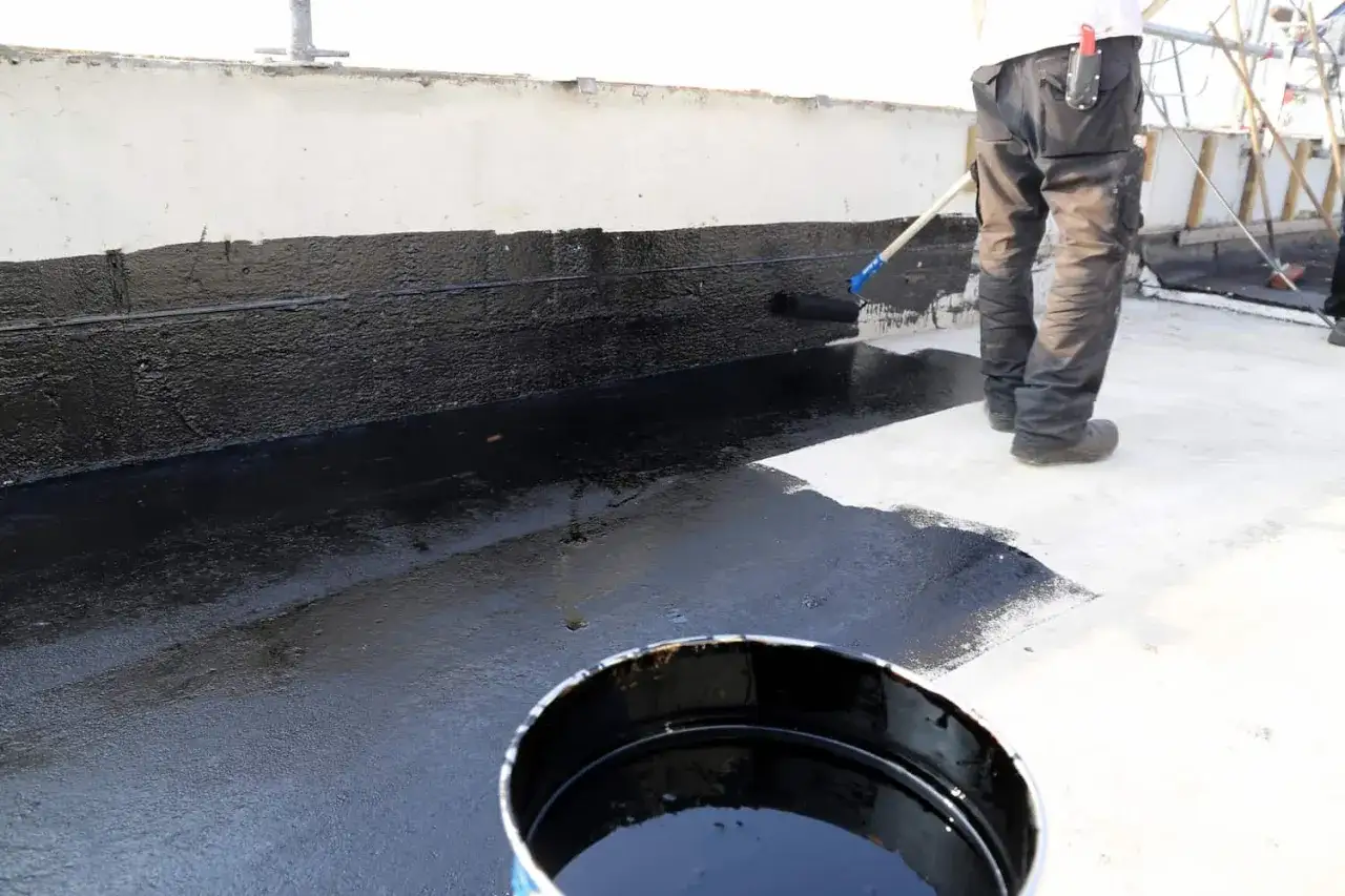 Arbeiter trägt mit Rolle mem bitumen voranstrich auf eine Wand auf. Ein Eimer mit schwarzer Flüssigkeit steht daneben.