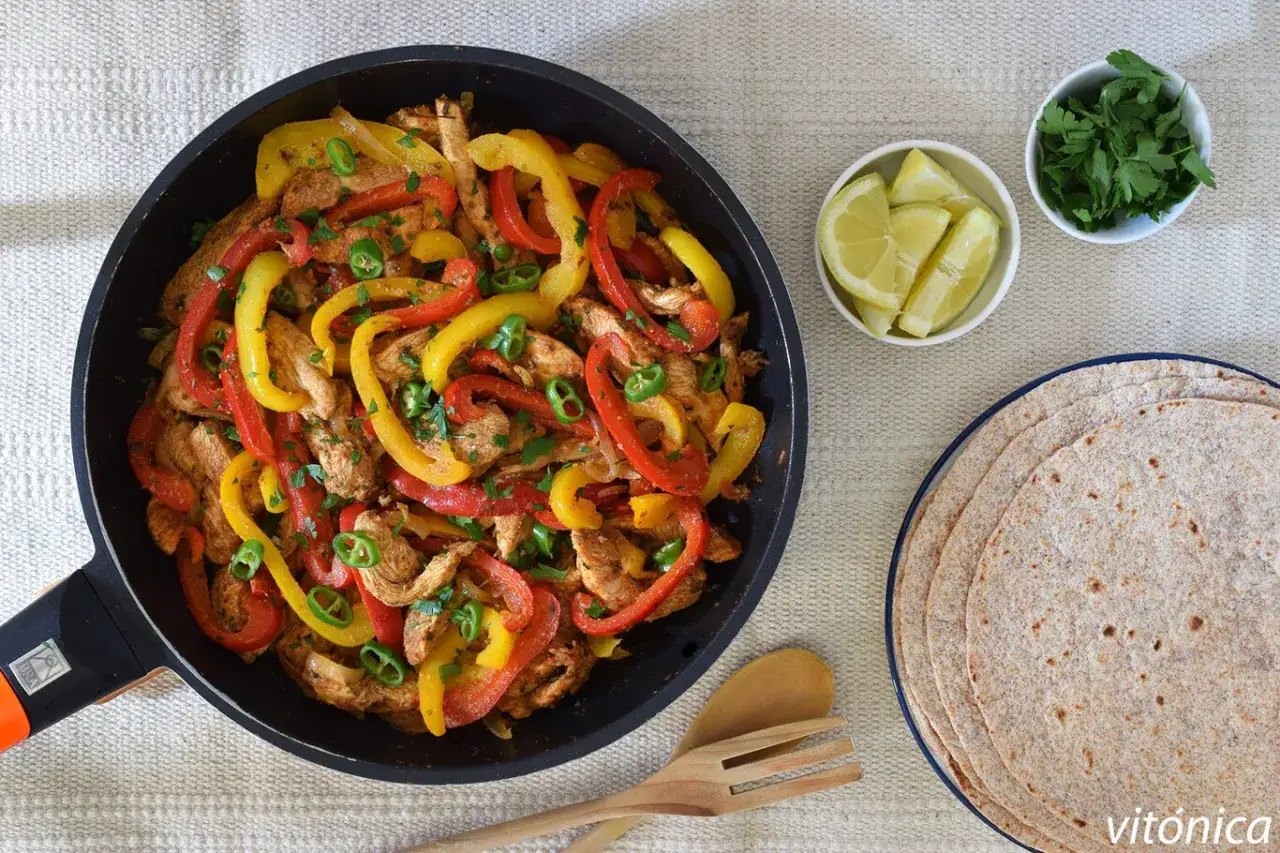 Fajitas de pollo con pimientos de colores, cebolla y chile verde, servidas en sartén con tortillas, limón y cilantro.