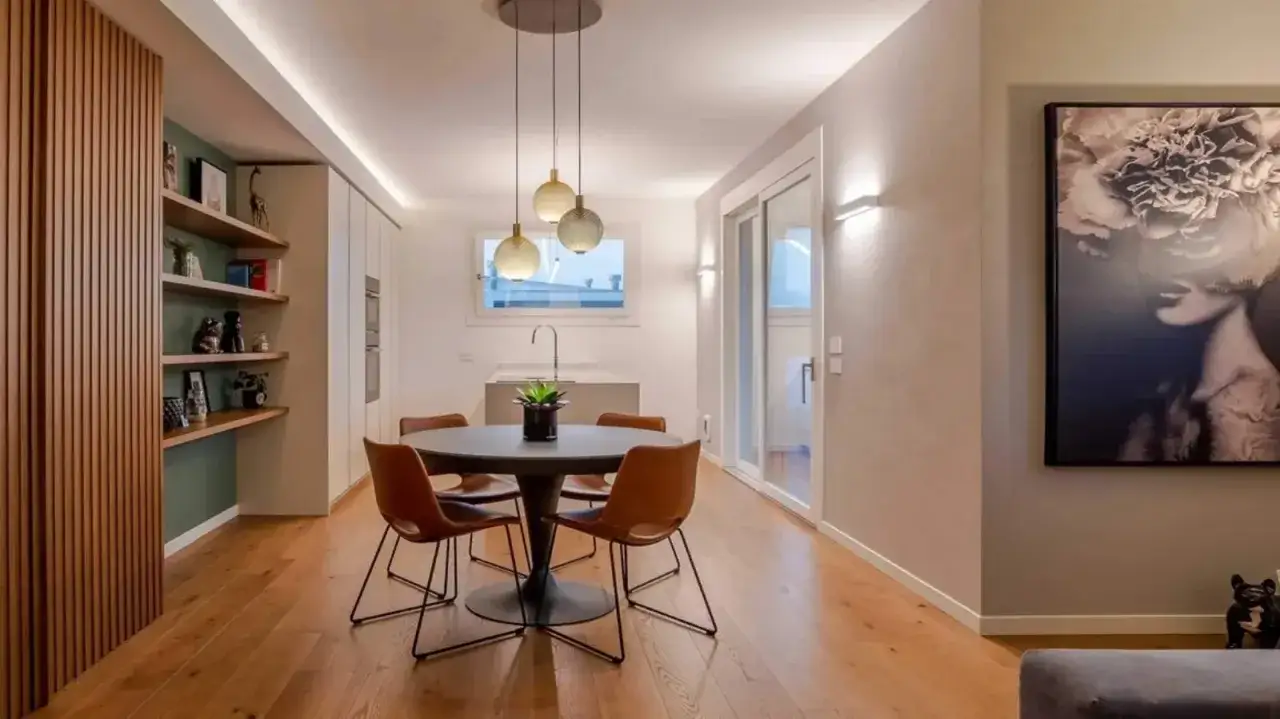 Tavolo rotondo con sedie in pelle, illuminato da lampade a sospensione. Parete con libreria e un quadro moderno. La luce più naturale valorizza l'ambiente.