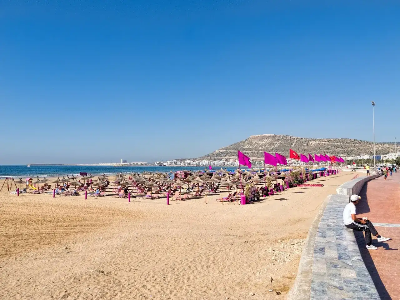 Plaża w Agadirze tętni życiem, mnóstwo leżaków i parasoli. W tle widać miasto i wzgórze. Czy Agadir jest bezpieczny? Wygląda na spokojne miejsce na wypoczynek.
