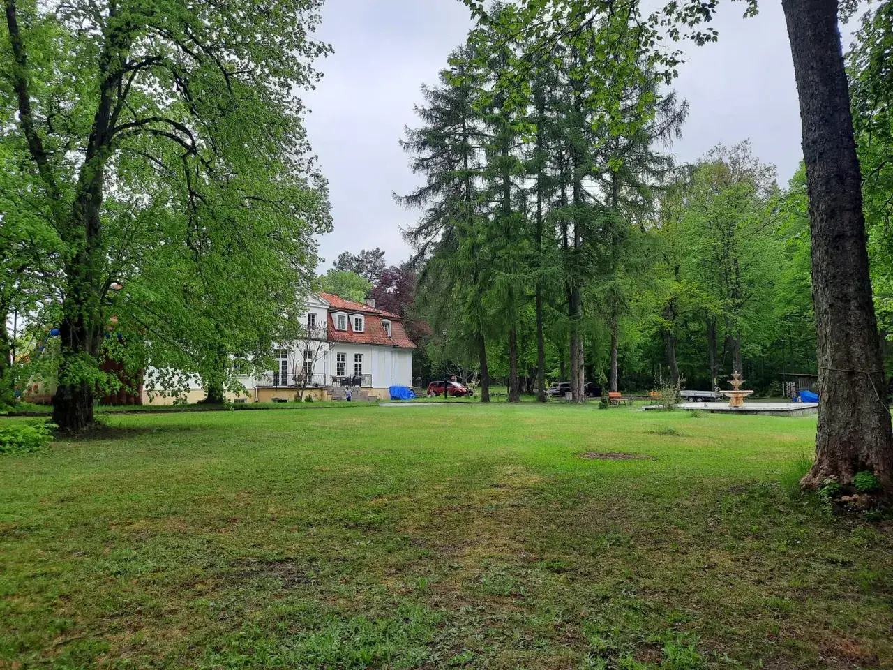 Biały dworek z czerwonym dachem otoczony zielenią. Na trawniku fontanna, drzewa i ławka. Miejsce jak z bajki, idealne na odpoczynek w **gierłoż**.