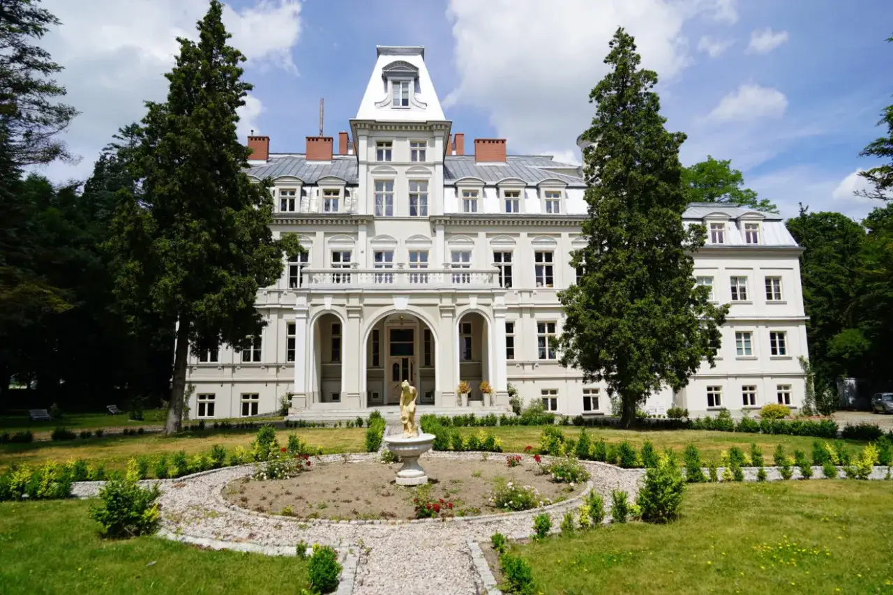 Elegancki pałac z białymi ścianami i licznymi oknami, otoczony zielenią. Przed nim fontanna z rzeźbą.