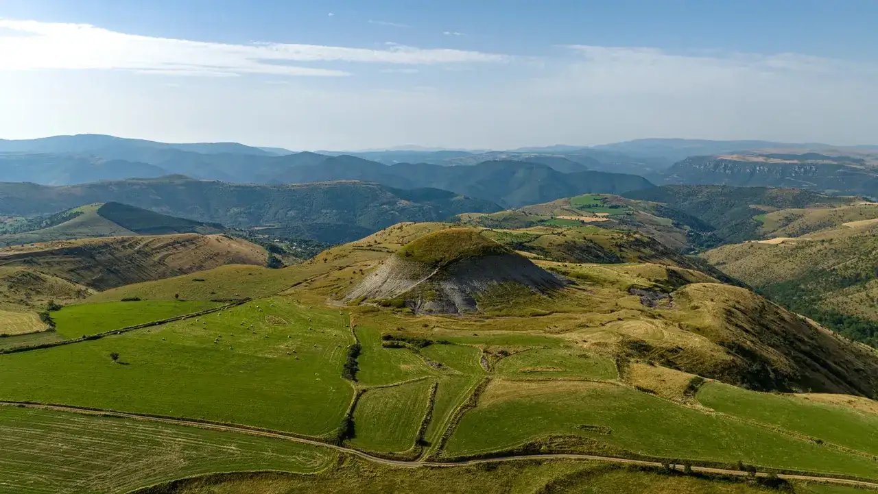 Paysage vallonné verdoyant, avec un cône volcanique au centre. Un chemin serpente à travers les champs, rappelant le chemin de Regordane.