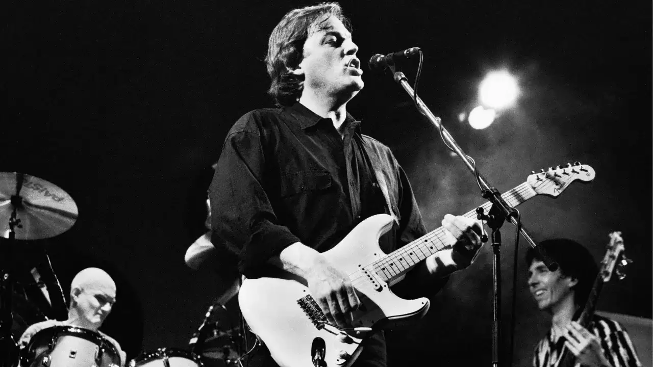 Un guitariste chante sur scène, dans un style musical rappelant Pink Floyd. Un batteur chauve est visible à gauche.