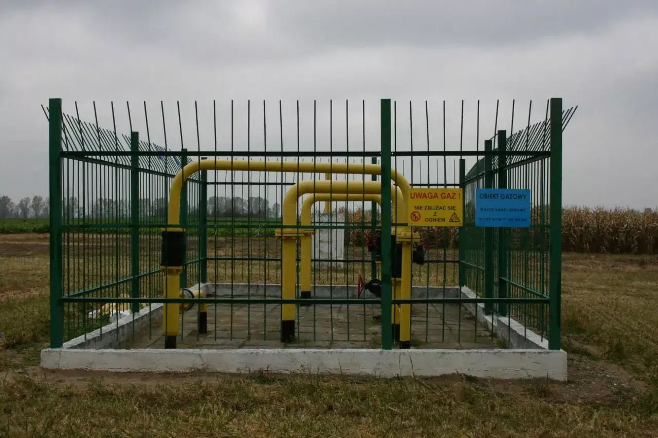 Żółty gazociąg wysokiego ciśnienia na działce, otoczony zielonym ogrodzeniem. Tabliczki ostrzegają przed gazem i ogniem.