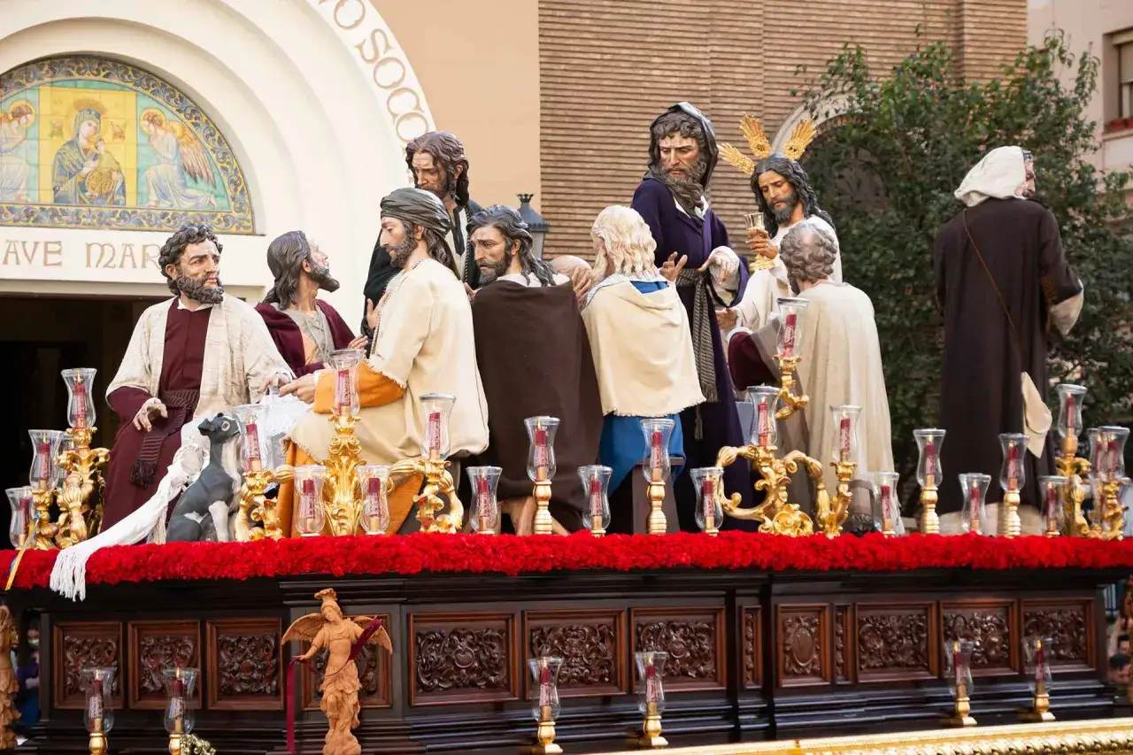 Escena de la Última Cena en una procesión de Jueves Santo en Zaragoza. Figuras religiosas y candelabros dorados sobre un paso decorado.