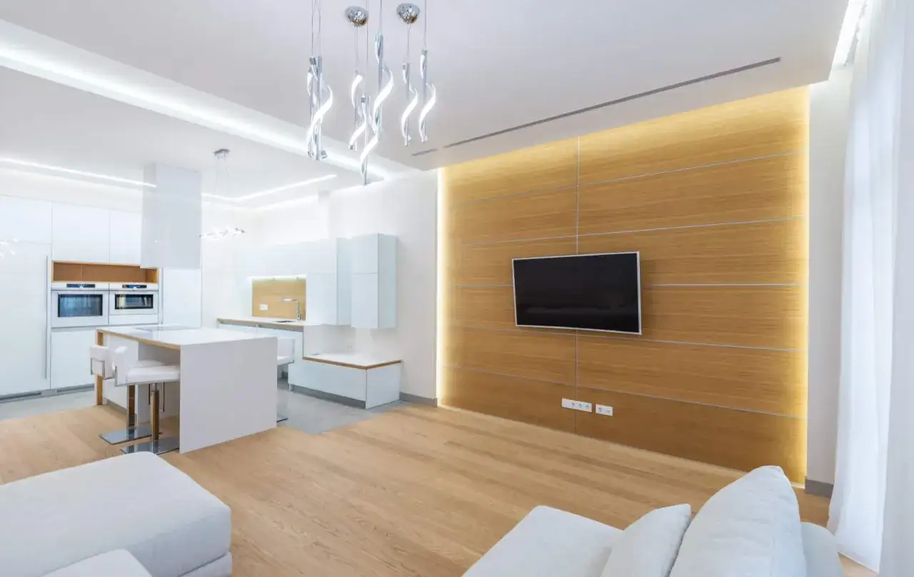 Cocina y sala de estar modernas con iluminación ambiental que realza los **lúmenes por m2**. Muebles blancos, isla de cocina y pared de madera con TV.