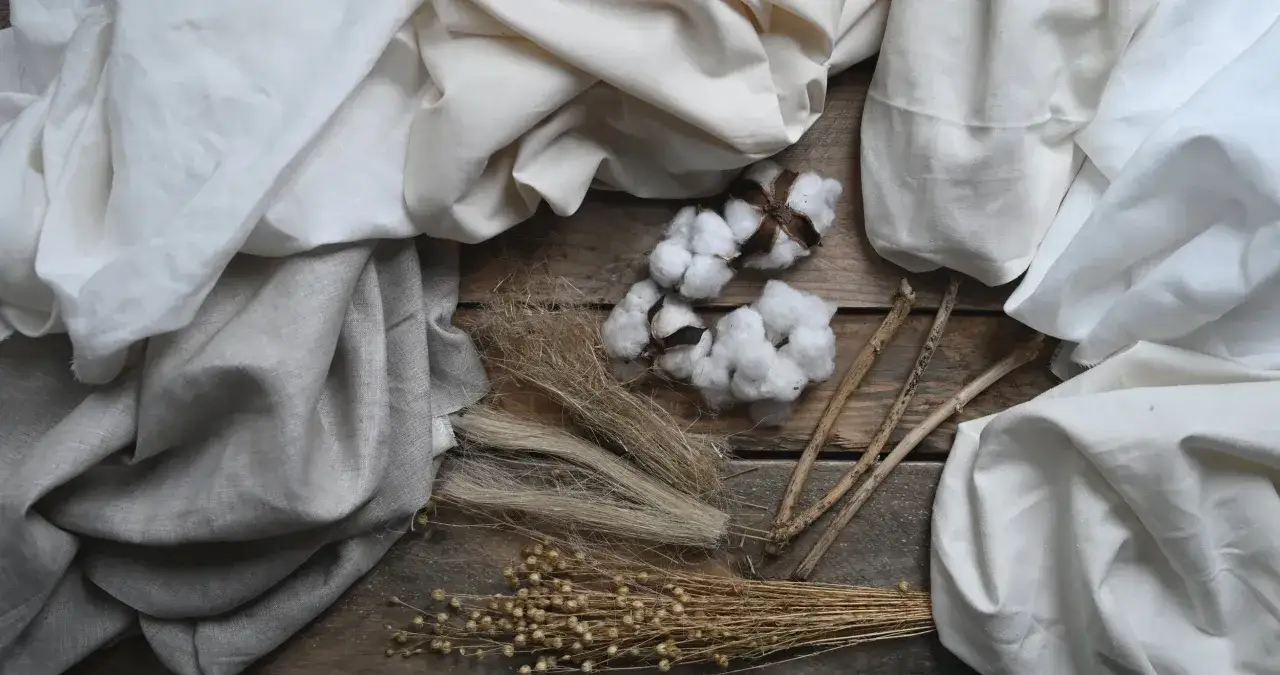 Tessuti naturali e fibre vegetali: cotone soffice, lino grezzo e steli di lino secco su un tavolo di legno.