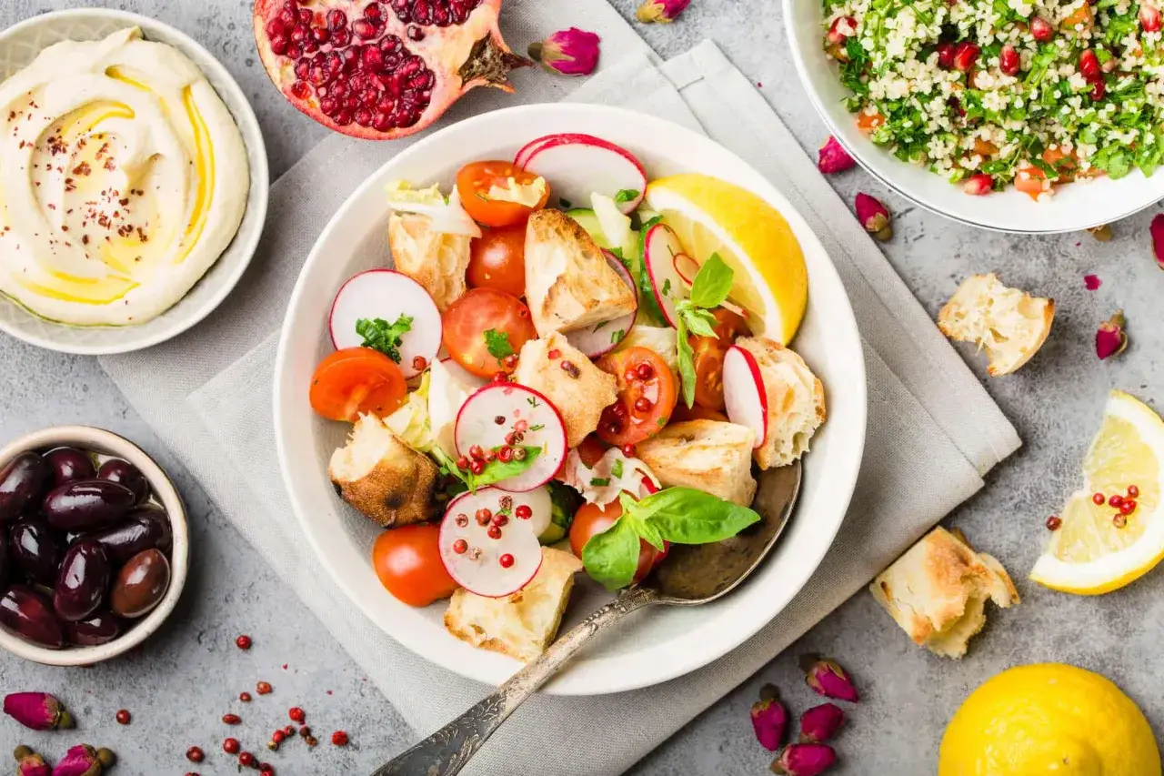 Insalata colorata con pane, pomodori, ravanelli e limone, perfetta per una dieta a zona equilibrata. Hummus, olive e melograno completano il pasto.