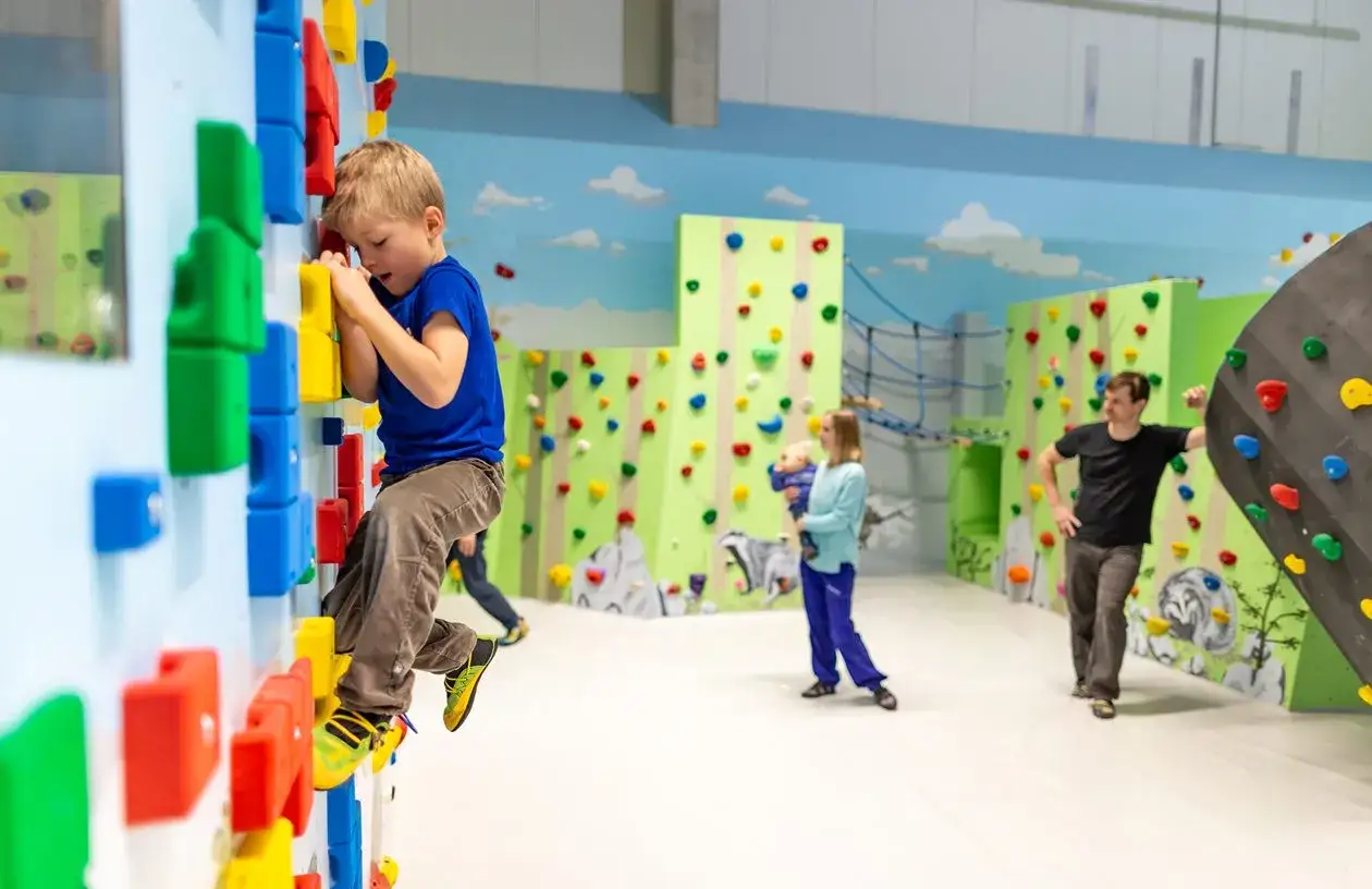 Ein kleiner Junge klettert konzentriert an einer bunten Wand. Im Hintergrund klettern weitere Kinder und Erwachsene. Ein toller Tag zum **klettern mit Kindern**!