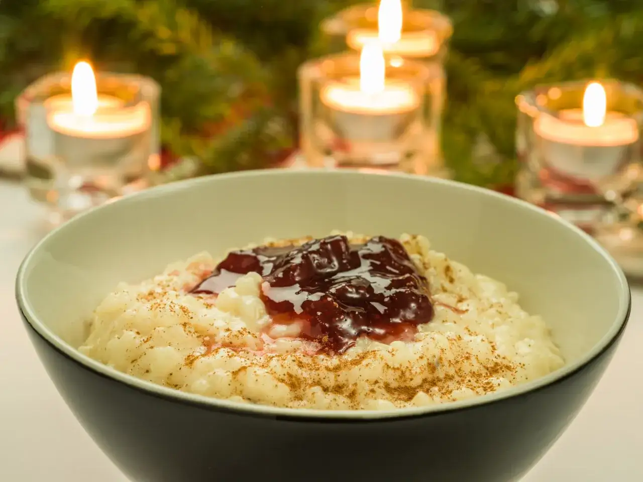 En sk&aring;l med kr&auml;mig risgrynsgr&ouml;t, toppad med sylt och kanel. T&auml;nda ljus och granris skapar en mysig julst&auml;mning, perfekt f&ouml;r ett risgrynspudding recept.