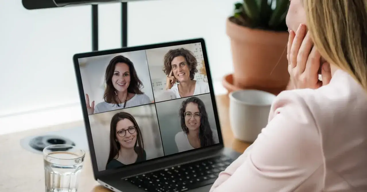 A woman participates in a digital coaching session with four smiling women on her laptop screen.