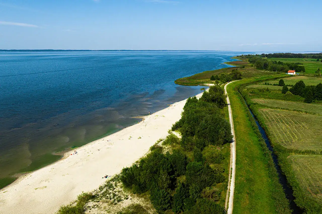 Słoneczna, piaszczysta plaża nad spokojnym jeziorem, otoczona zielenią i polami. Idealne miejsce na wypoczynek.