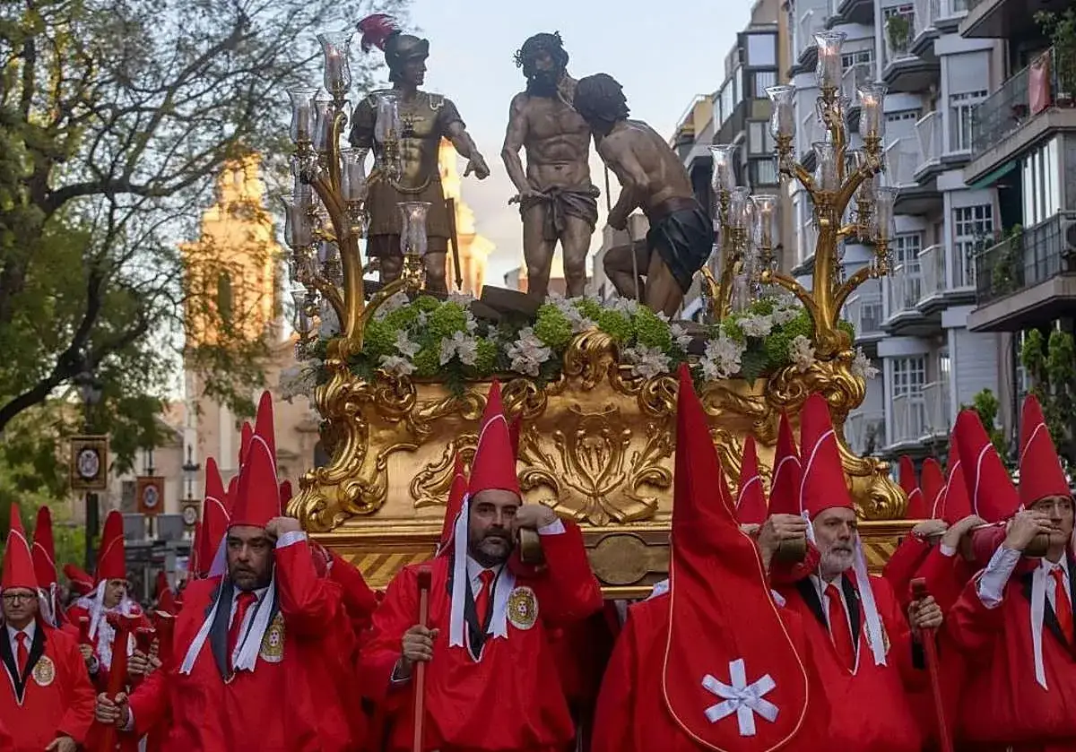 Hombres con túnicas rojas y capirotes participan en una procesión de Miércoles Santo, portando un paso dorado con figuras religiosas.