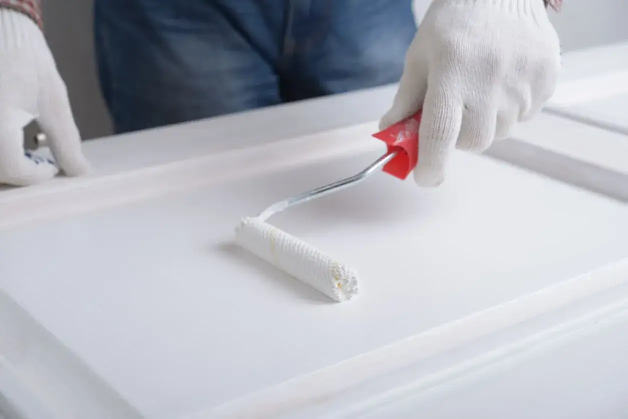 Manos enguantadas pintando puertas de madera con rodillo blanco.