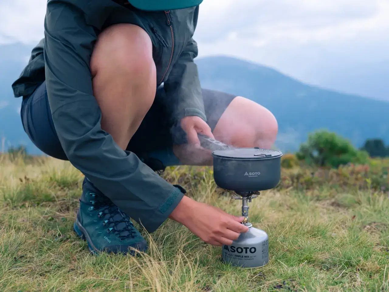 Person kocht mit einem Soto Windmaster Kocher in den Bergen. Dampf steigt aus dem Topf auf.