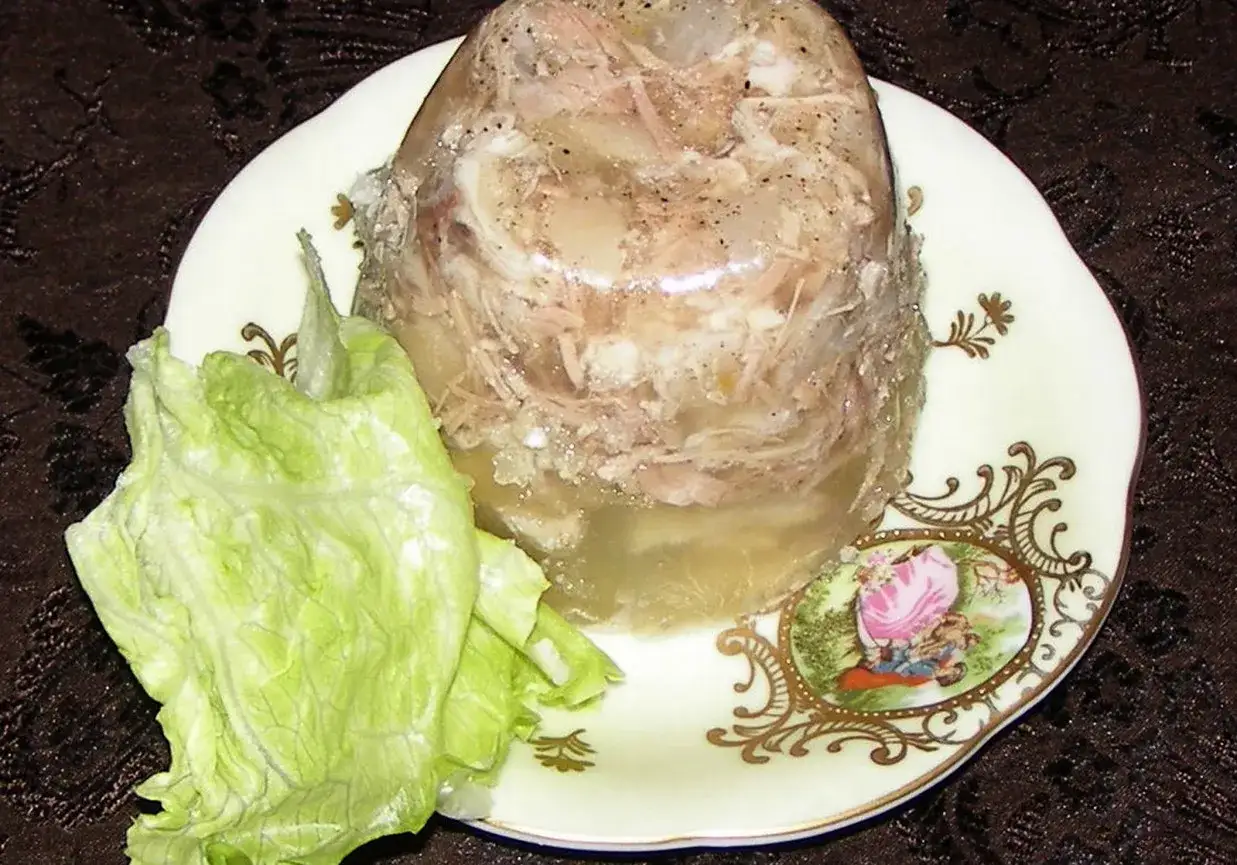 Pyszna galareta z golonki, z kawałkami mięsa w galaretce, podana z liściem sałaty na ozdobnym talerzu.