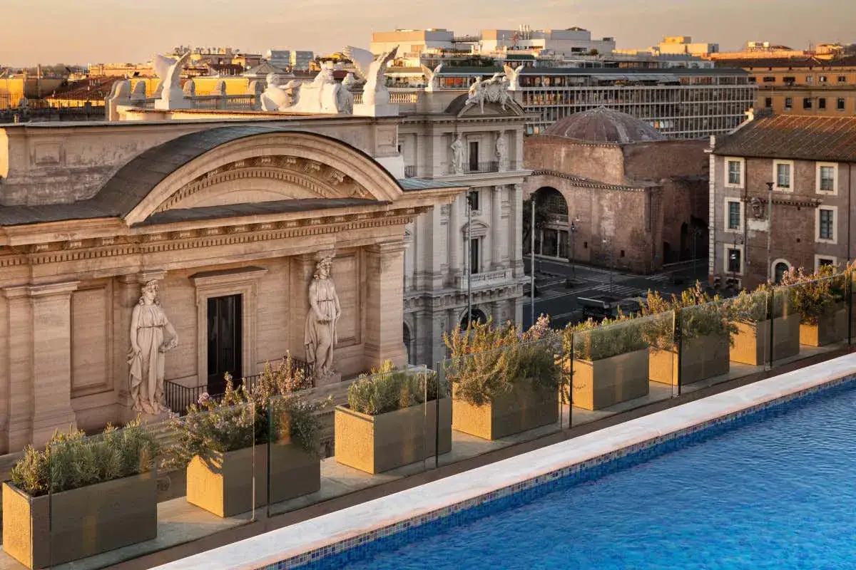 Vista panoramica dal tetto dell'Anantara Palazzo Naiadi Rome Hotel, con piscina, piante in vaso e antichi edifici romani al tramonto.